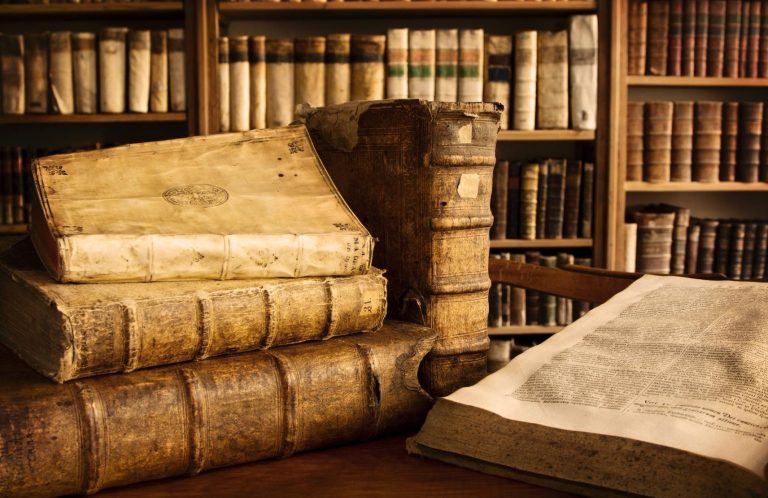 Ein Haufen alter Bücher auf einem Tisch, mit Bücherregalen im Hintergrund.