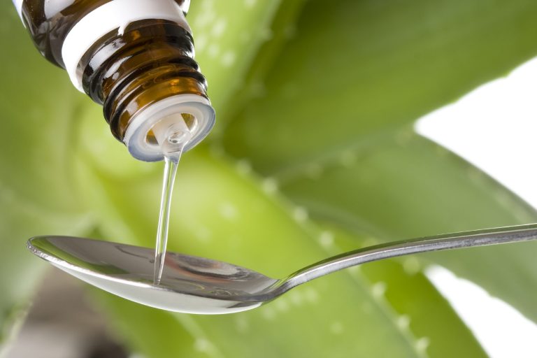 Braunflasche gießt Flüssigkeit auf einen Löffel, umgeben von grünen Aloe-Vera-Blättern.