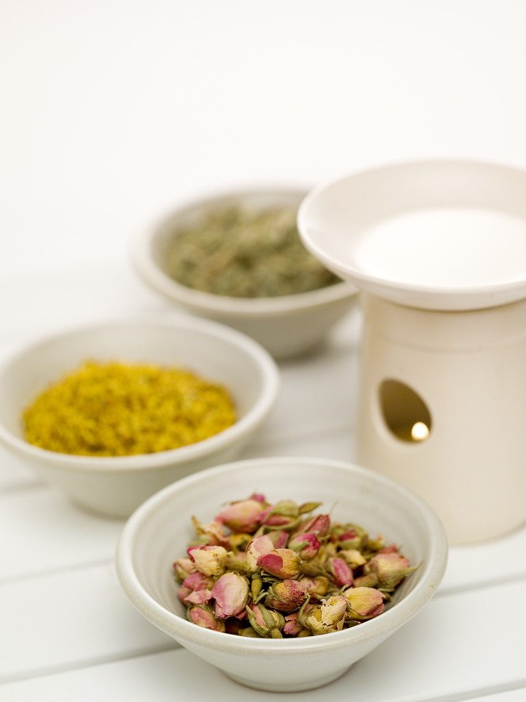 Tropfen von Gewürzen und getrockneten Rosenblüten in Schalen neben einer Aromalampe.