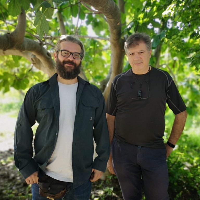 Zwei Männer stehen vor einem Baum, lächeln und betrachten die Kamera.