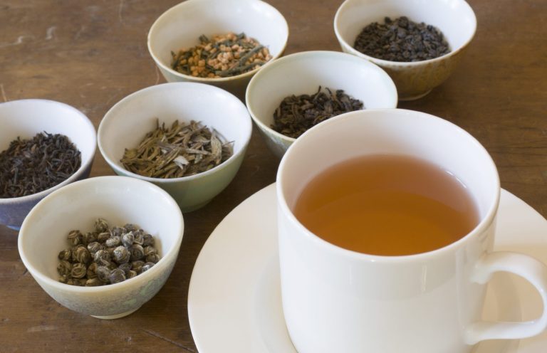 Tasse Tee vor mehreren Schalen mit verschiedenen Teekräutern und Gewürzen.